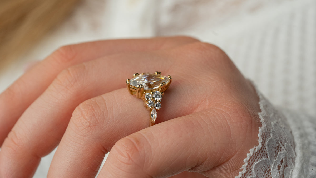 A close-up of a hand wearing a yellow gold ring with a marquise-cut clear center stone and smaller accent stones, set against a soft white lace sleeve and blurred skin tones.