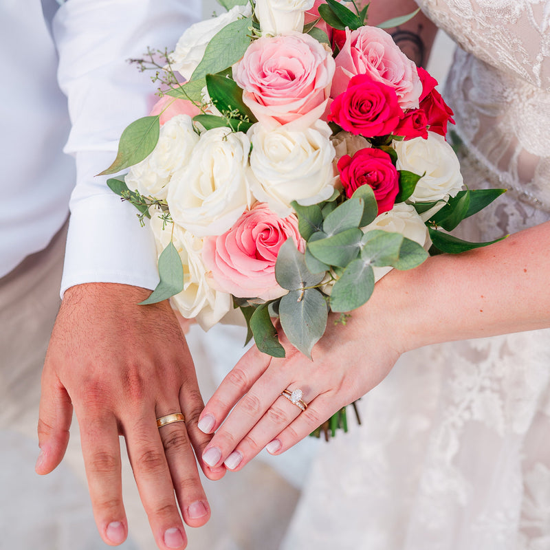 Newlywed couple showing his and hers 14K yellow gold wedding rings on hands with floral bouquet