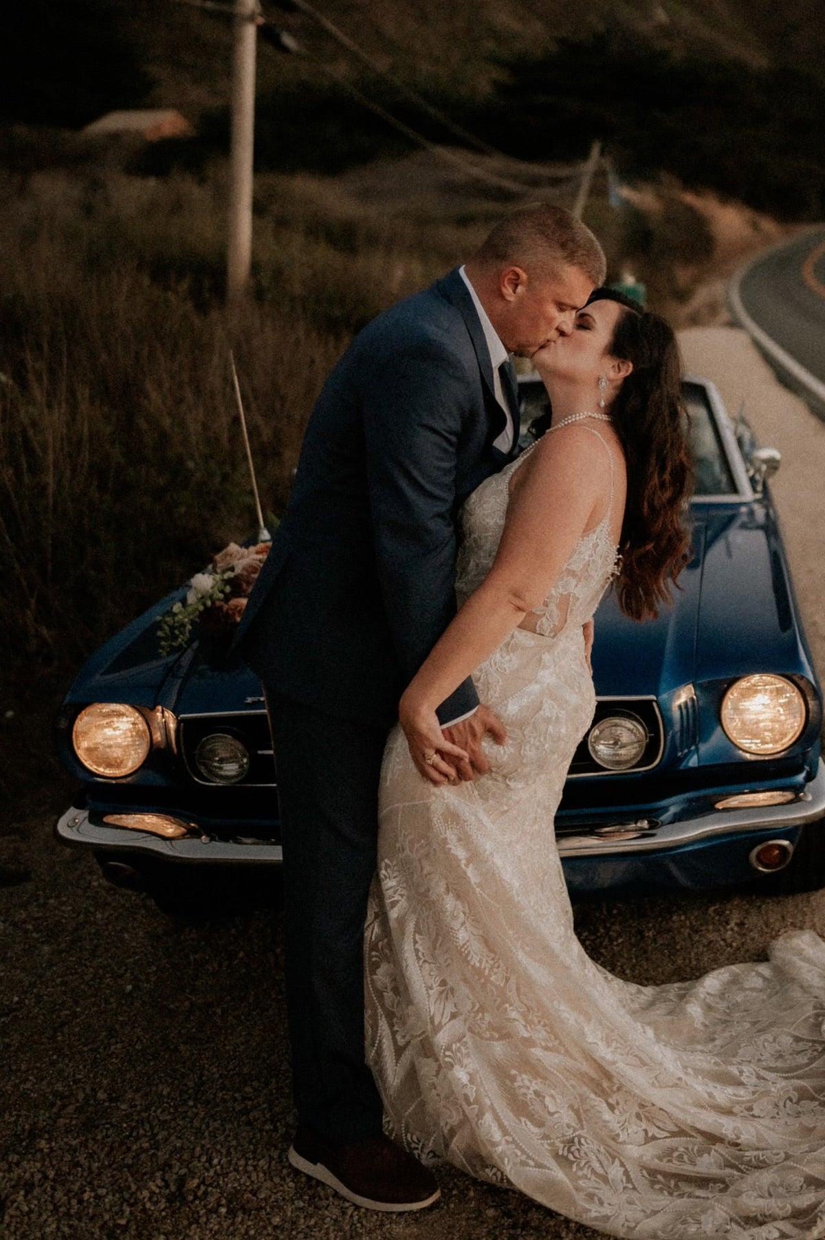 Bride and groom embracing in front of a vintage car on a road with mountains in the background