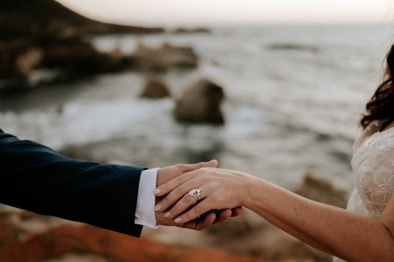 Two people holding hands showing her custom wedding set with a blurred natural background