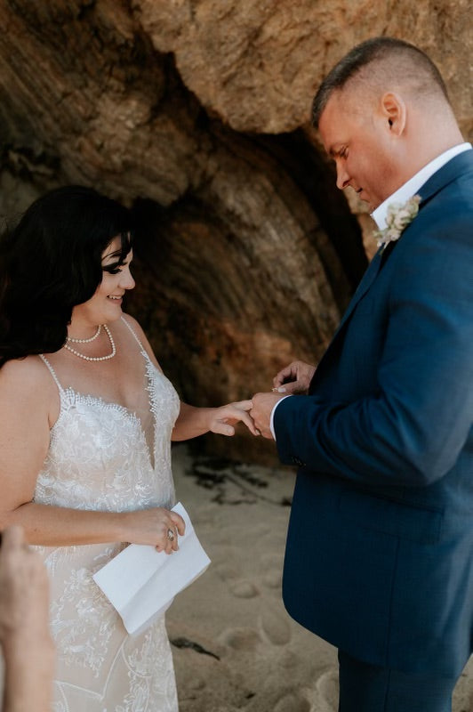 Bride and groom in wedding attire exchanging rings on a beach with a natural rock formation in the background.