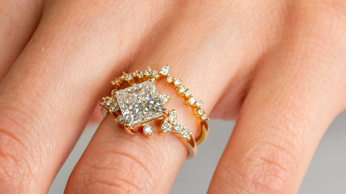 Constellation engagement ring on the hand: the Zakari with a princess cut diamond in yellow gold, stacked with the Twilight curved scatter diamond wedding ring